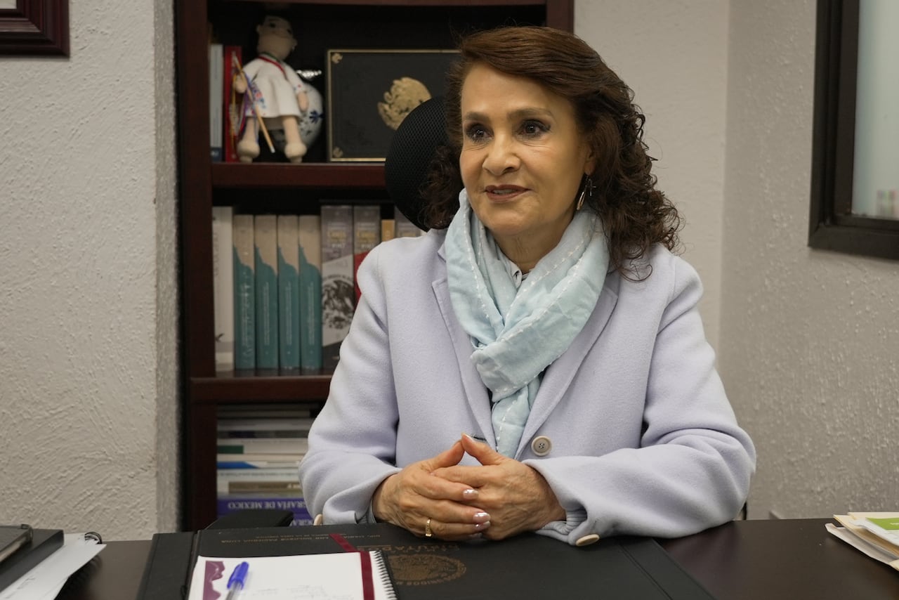 A woman sits at a desk with her hands joined in front of her.