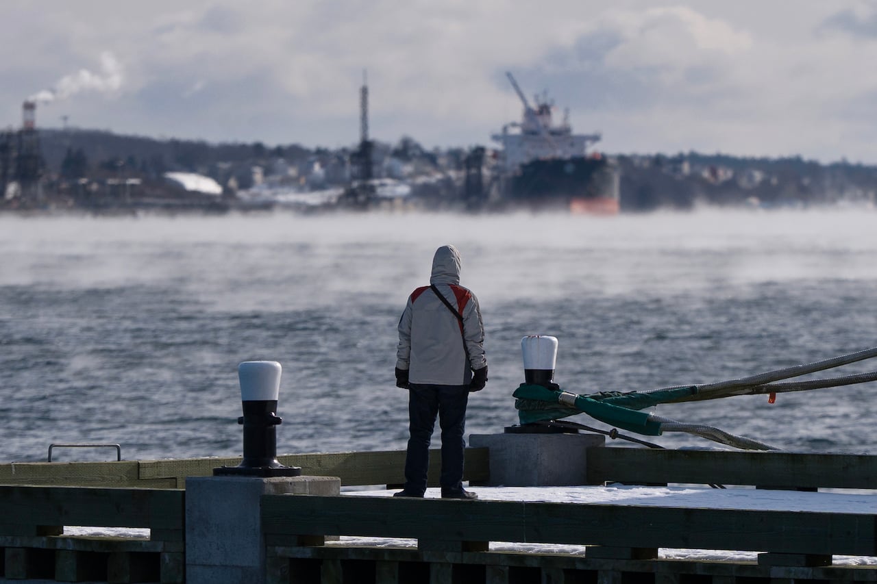 A person in winter clothes stands at a pier as fog emanates from the water.