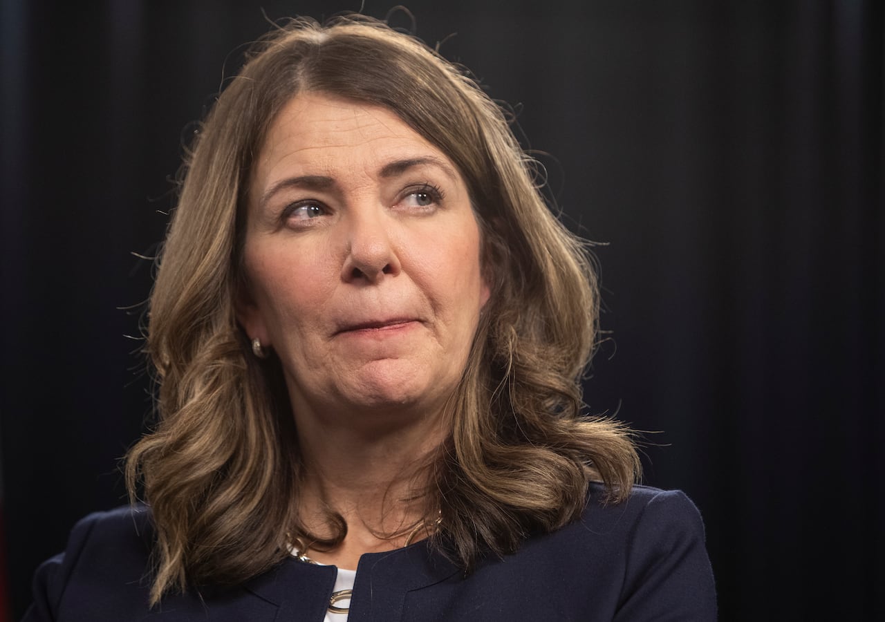 A woman with wavy brown hair stands in front of a navy blue background.