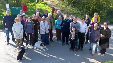 A group of 24 villagers of all ages and two dogs are gathered in North Moreton in front of a grass verge