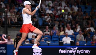 Tenisistka podczas meczu, uderza piłkę rakietą na korcie, w tle trybuny z widzami.