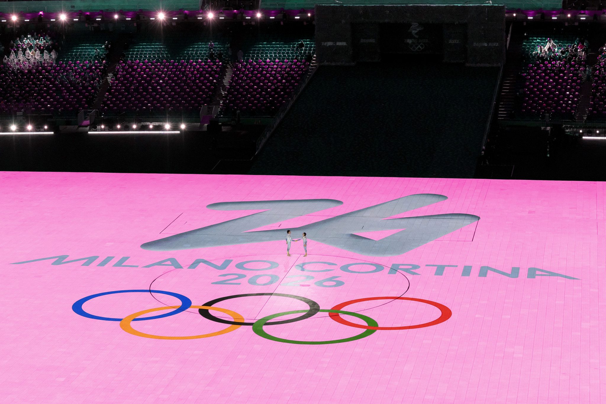 Cérémonie avec logo de Milano Cortina 2026 et anneaux olympiques sur fond rose.