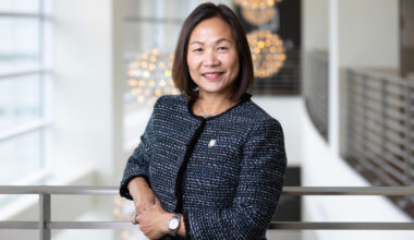 UNO Chancellor Joanne Li poses for a photo. She is wearing a black blazer and leaning against a railing with light fixtures in the background.