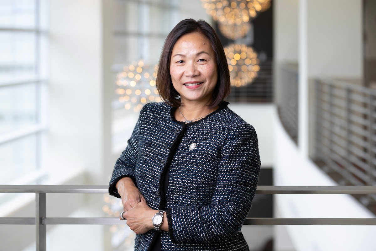 UNO Chancellor Joanne Li poses for a photo. She is wearing a black blazer and leaning against a railing with light fixtures in the background.