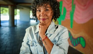 A woman with short curly hair stands resting her chin on her hand. Behind her are colorful murals.