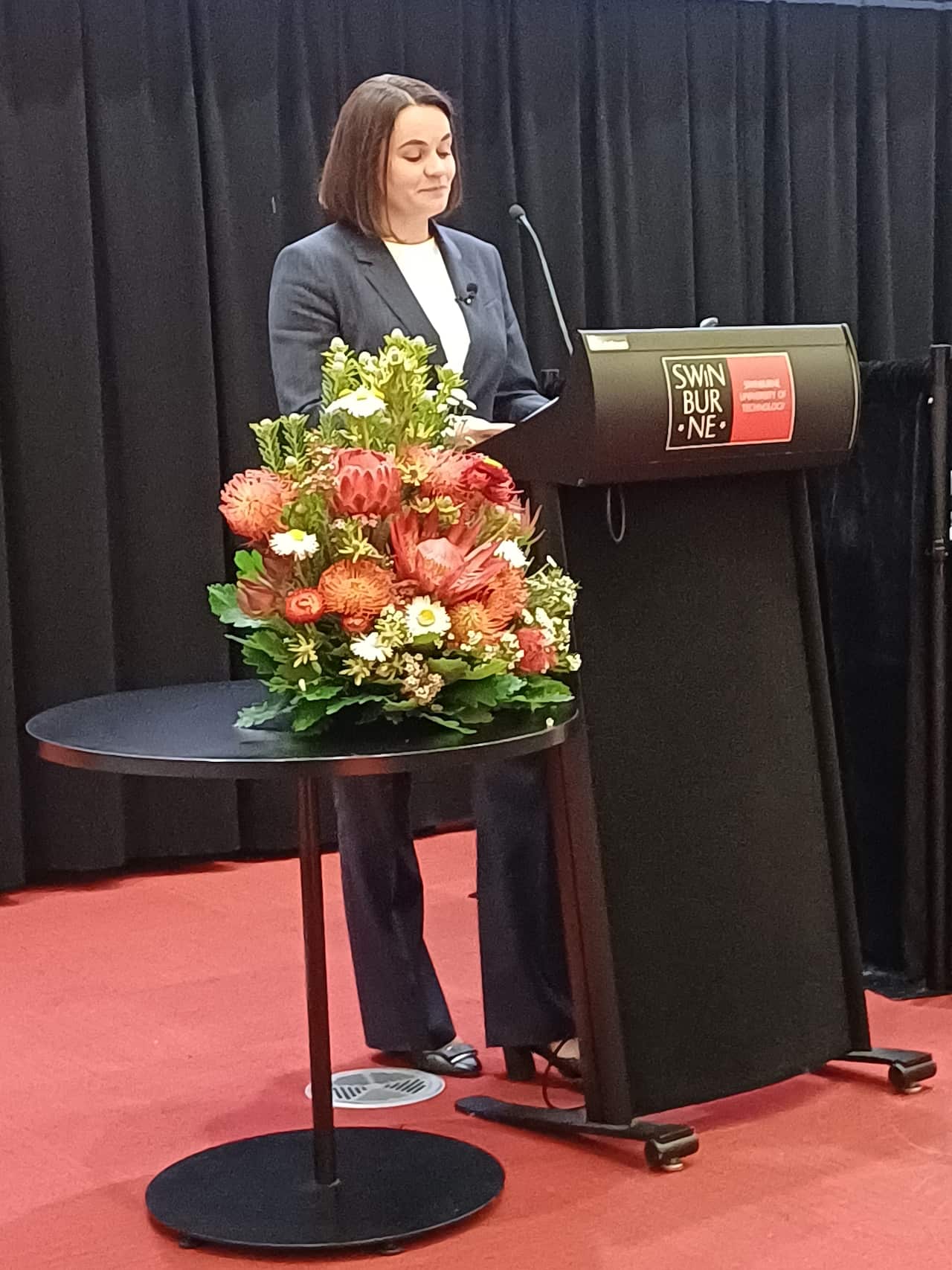 A woman stands behind a podium with a microphone, smiling as she speaks, in front of a black curtain, with a large floral arrangement on a small table beside the podium.