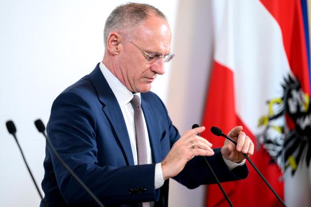 Ein Mann im Anzug (Innenminister Karner) richtet Mikrofone vor einer rot-weiß-roten Fahne mit einem Adler.