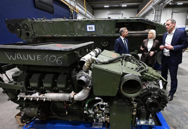 Drei Personen stehen in einer Werkhalle neben einem ausgebauten Militärfahrzeugmotor und einem gepanzerten Fahrzeug.