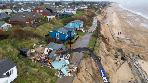 PA Media A bulldozer rips apart a clifftop chalet, with the debris of windows and wooden partitions scattered on the ground. Three other similar properties stand either side, with rows of other houses behind. To the right of the image, a sandy beach and shoreline stretches off into the distance.  