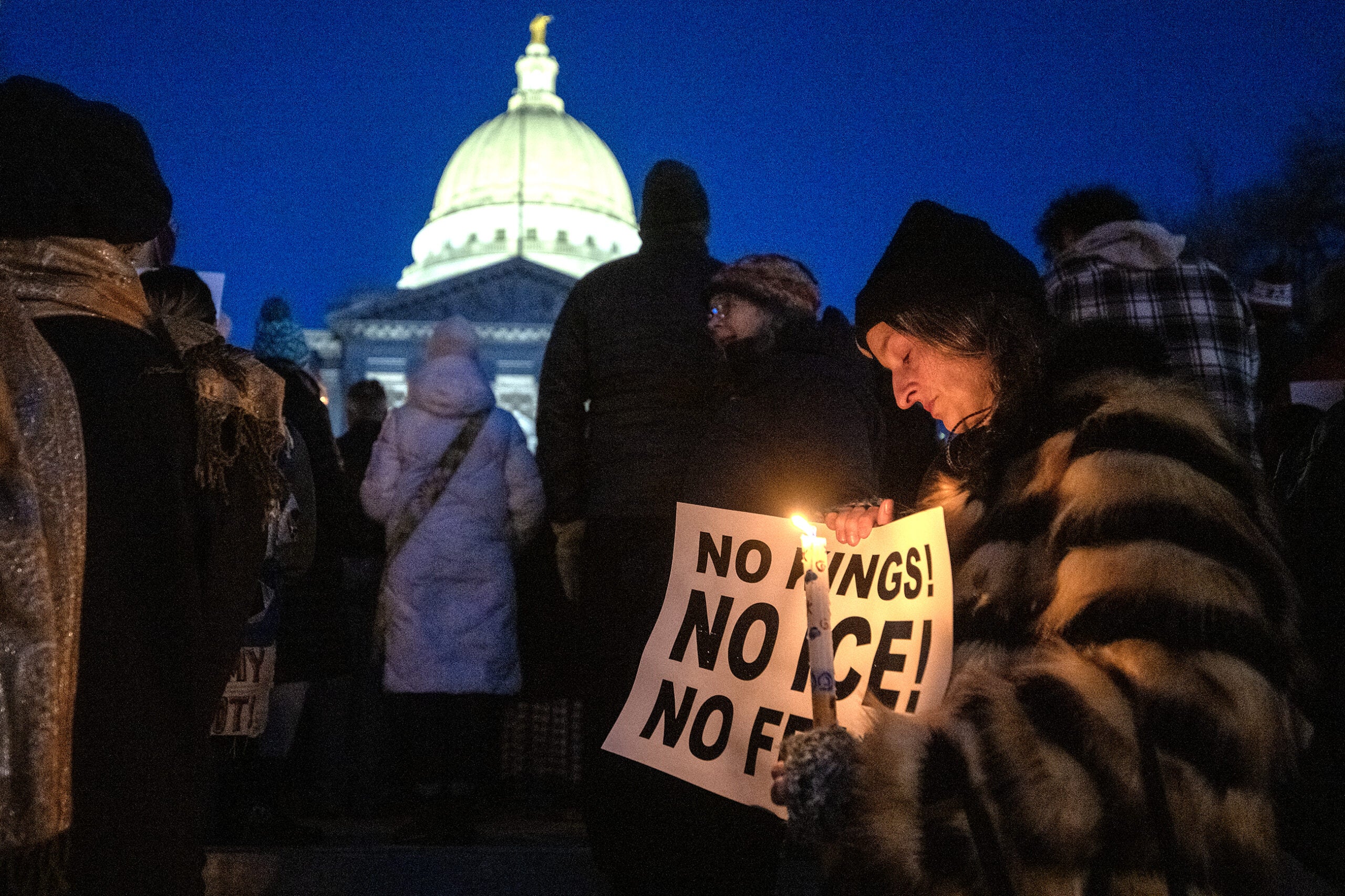 A person holding a candle and a protest sign reading NO BANS! NO ICE! NO FEAR! stands among a crowd outside a domed government building at night.