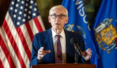 An older man in a suit and tie speaks at a podium with a microphone, with American and Wisconsin state flags in the background.