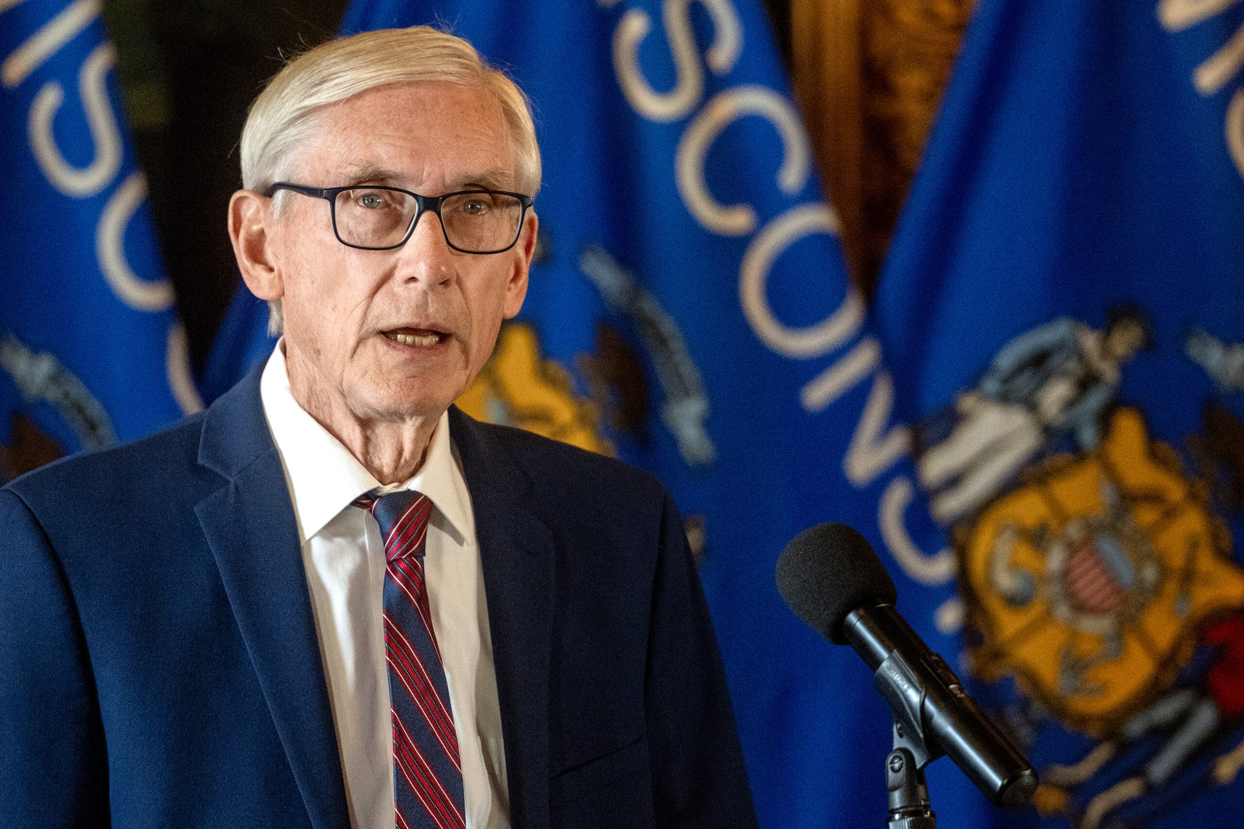 An older man in a suit and glasses speaks at a microphone, with Wisconsin state flags visible in the background.