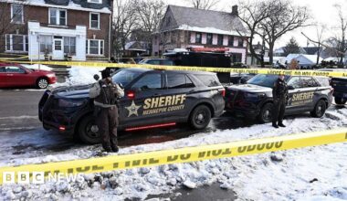 Police tape is shown blocking off a snow-covered residential street. Two sheriff cars are in the foreground with officers standing in front of them.