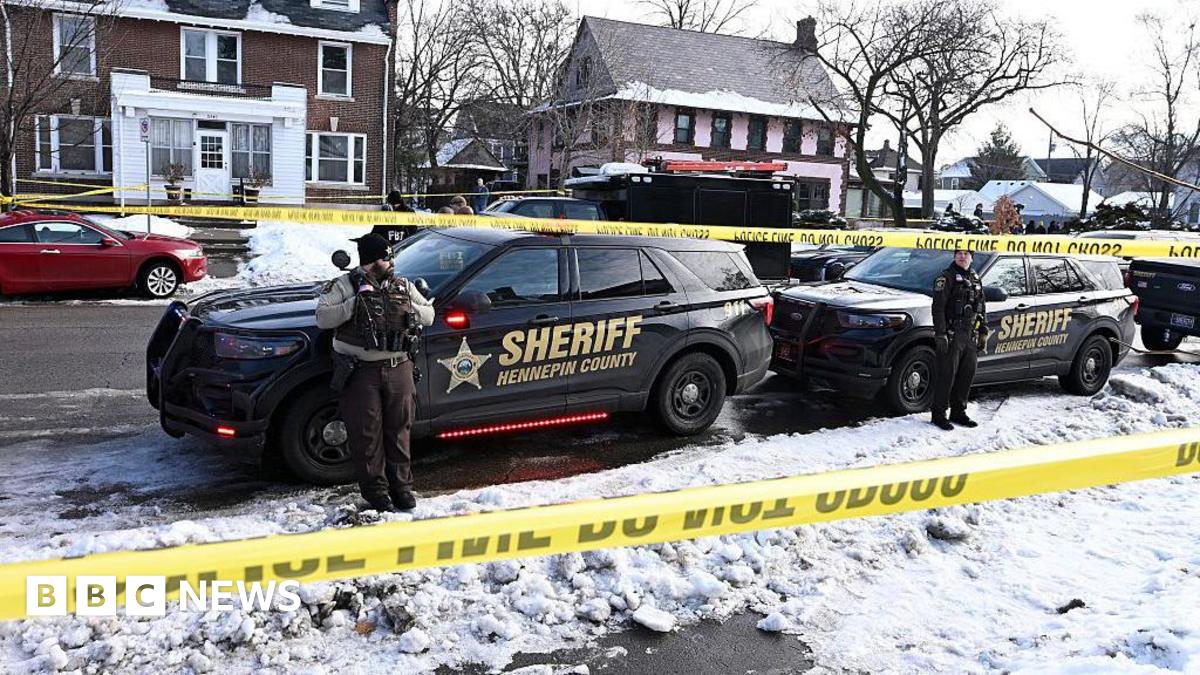 Police tape is shown blocking off a snow-covered residential street. Two sheriff cars are in the foreground with officers standing in front of them.