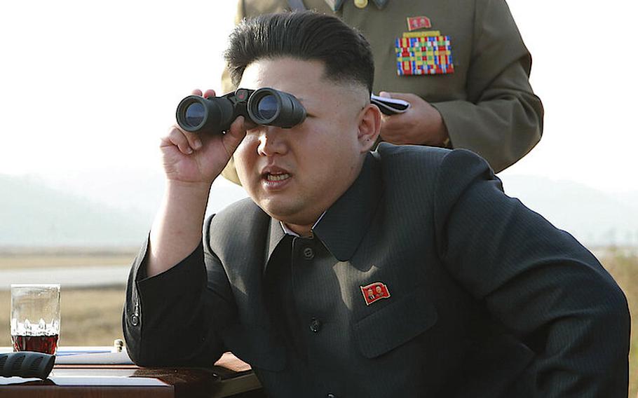 Kim Jong Un, wearing a black collared shirt, rests his elbow on a table and holds a pair of binoculars to his eyes, while a military officer in dress uniform stands behind him.