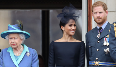 Queen Elizabeth wearing a blue coat standing next to Meghan Markle, in a navy dress and hat, and Prince Harry in a military uniform