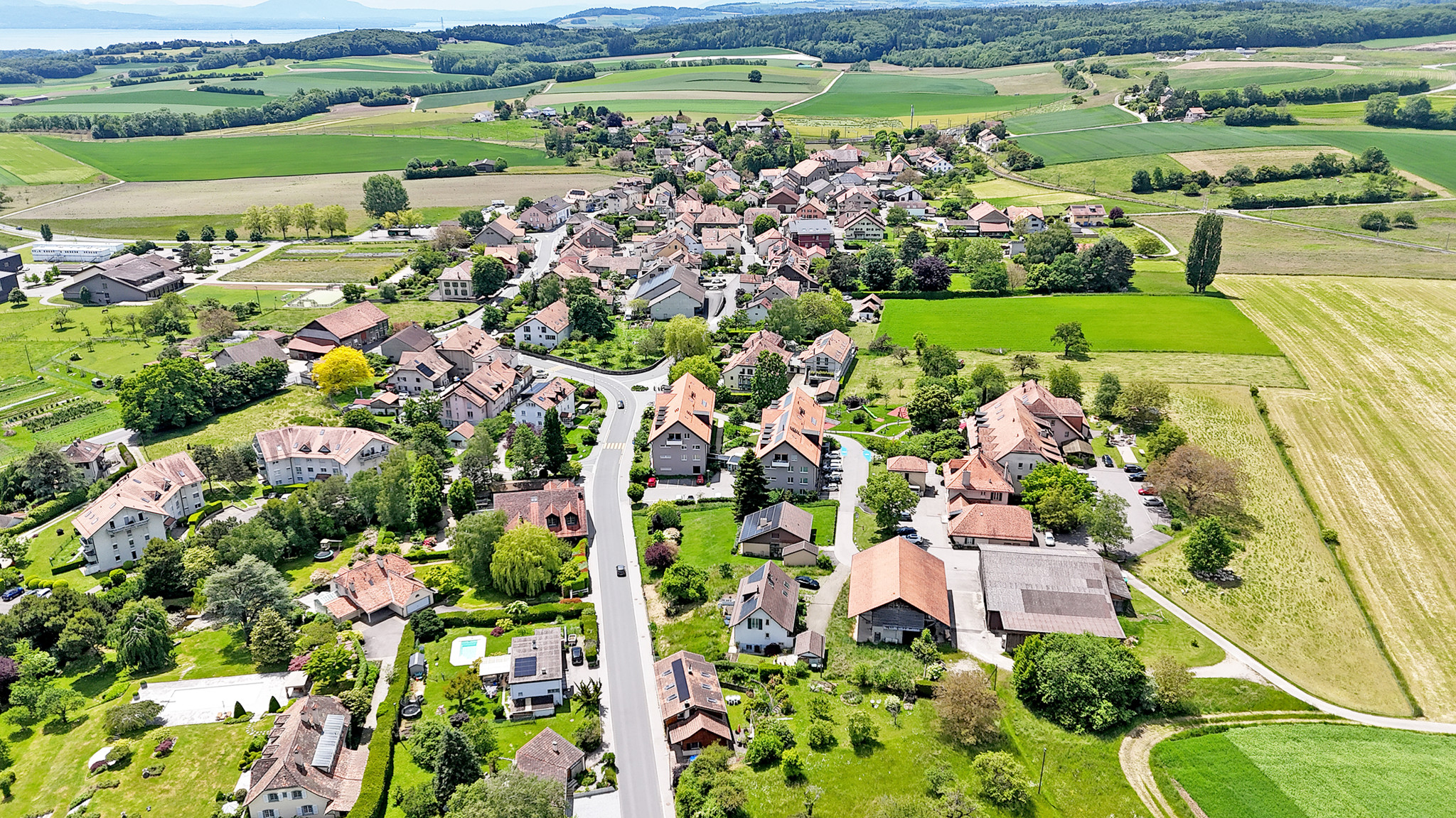 Vue aérienne d’un village suisse entouré de champs verdoyants, avec des maisons aux toits rouges et des routes serpentant à travers eux.
