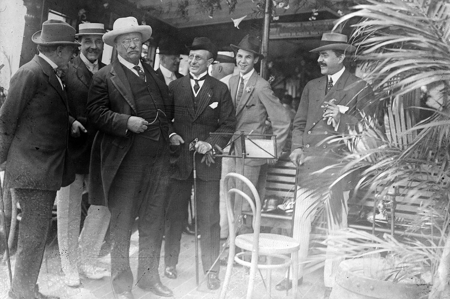 Teddy Roosevelt, waring a brimmed white hat, stands among men dressed in typical suits and hats of the period. A plant with palm leaves frames the picture at right. A cane-backed chair is in the foreground.