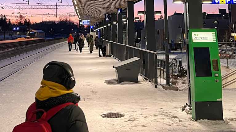A snowy train station platform with people waiting and an electronic board announcing train delays.
