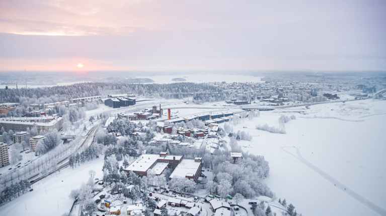 Aerial photo of a snowy city and natural landscape, with an orange sun seen low in the sky.