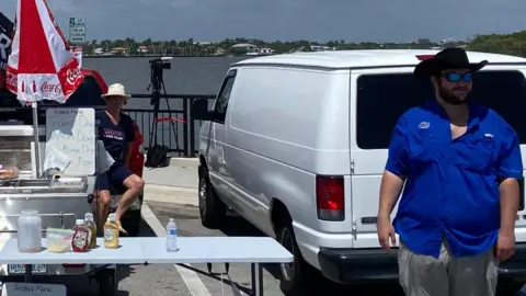 Bernd Debusmann Jr/BBC News Dirk Frazier, dressed in blue with a black cowboy hat, pictured on a bridge next to his hot dog stand with the waterways of Palm Beach in the background.