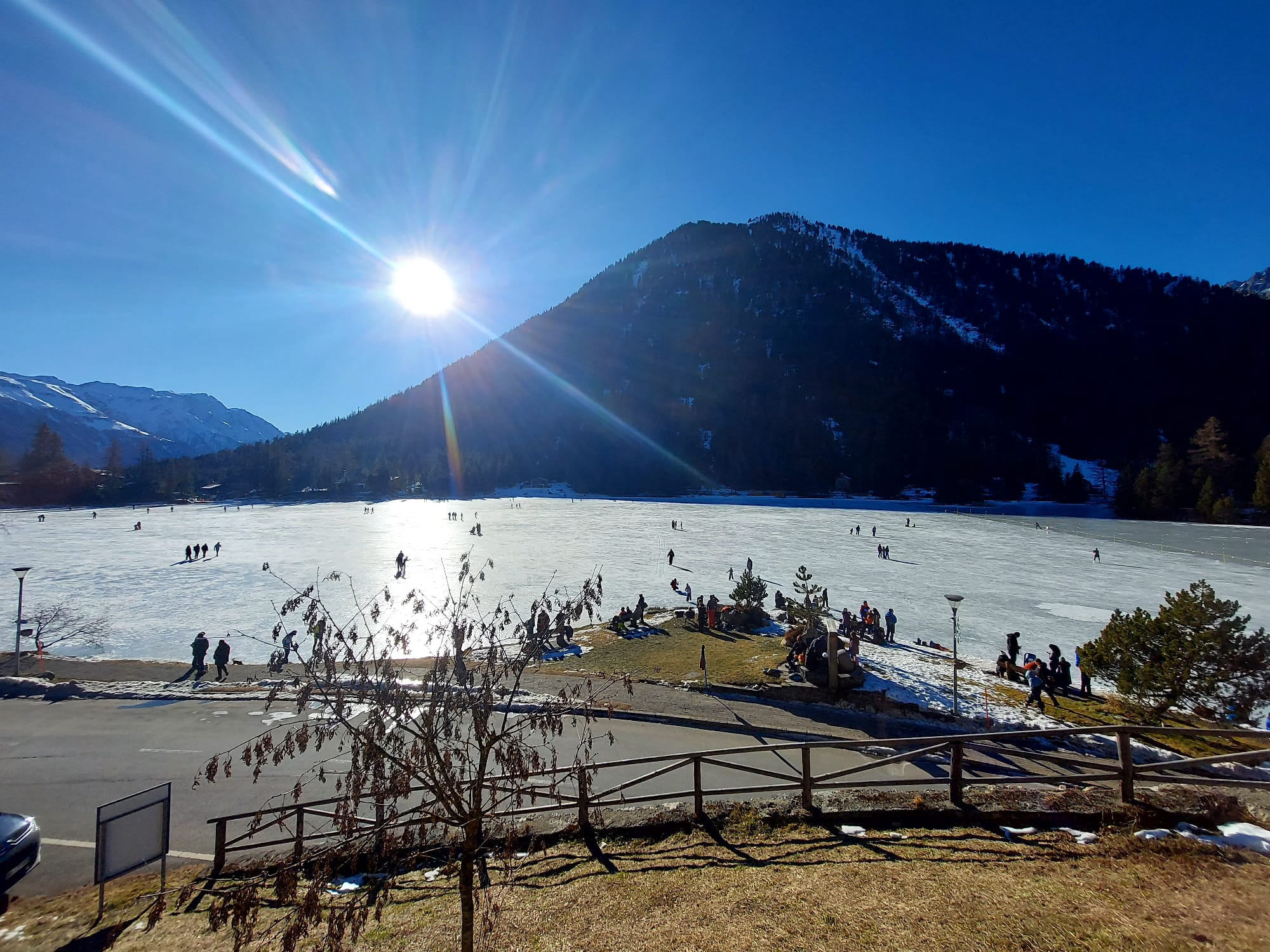 Paysage hivernal avec un lac gelé entouré de montagnes, des gens patinent et profitent du soleil éclatant sous un ciel bleu.