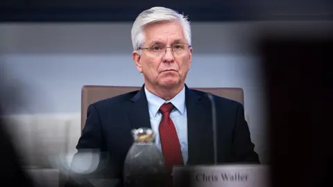 Bloomberg via Getty Images Christopher Waller, wearing a dark suit and red tie sits behind a table, with blurry placard bearing his name on it