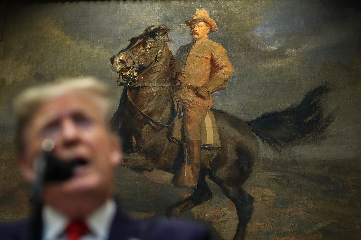 Donald Trump is seen out of focus in the foreground as he speaks into a microphone. In focus behind him is a painting of Teddy Roosevelt wearing safari gear astride a rearing horse.