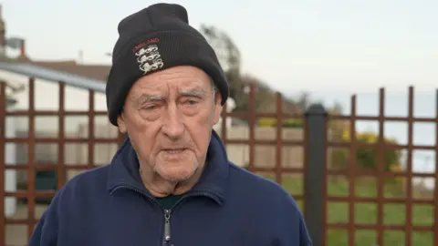 Qays Najm/BBC A man in a black woolly beanie hat with "England" and three lions on the front. He has grey stubble and is wearing a blue zipped-up fleece.