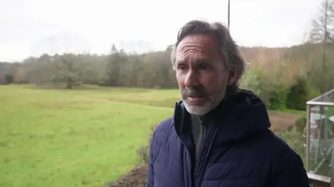 A man stands outside. Behind him is a field with trees. He is wearing a blue jacket and has grey slicked-back hair. 