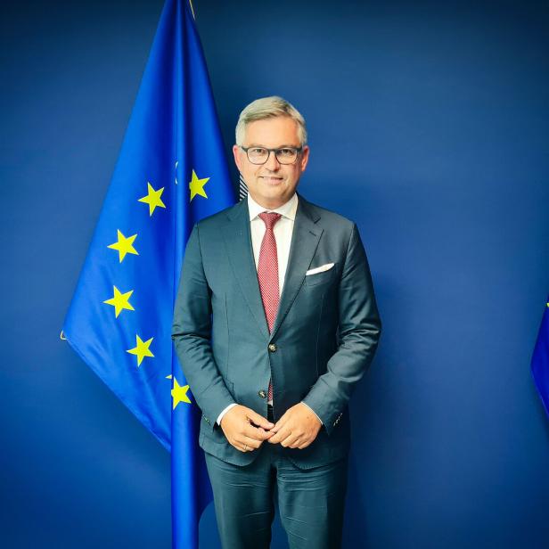 Migrationskommissar Magnus Brunner (ÖVP) steht vor einem blauen Hintergrund und zwei EU-Flaggen. Er ist am Rande der Plenarsitzung des Europäischen Parlaments Anfang September 2025 in Straßbourg.