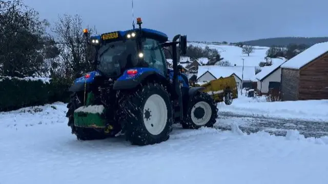 A blue tractor sits on snow covered ground, there is less snow where the road is.