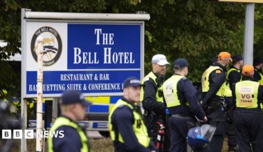 Police officers wearing caps and hi-vis jackets standing in front of a large blue sign that says "The Bell Hotel".