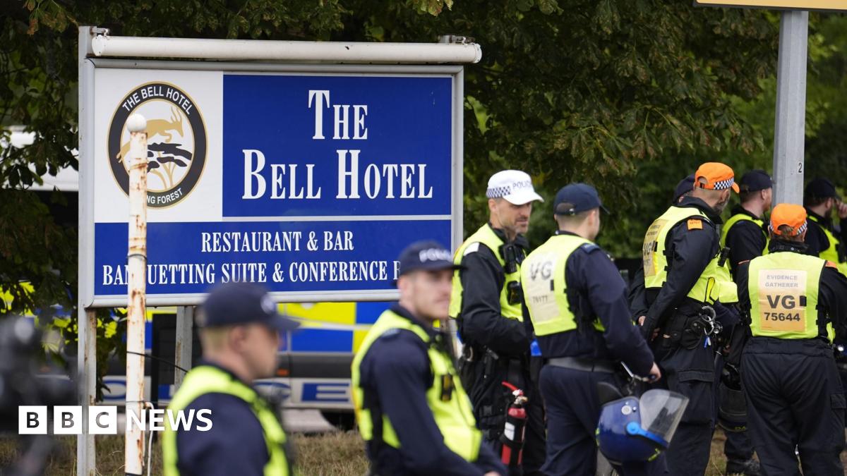 Police officers wearing caps and hi-vis jackets standing in front of a large blue sign that says "The Bell Hotel".