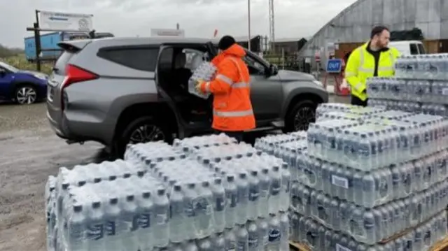 Bottles of water are unloaded at a distribution point by workers in hi vis.