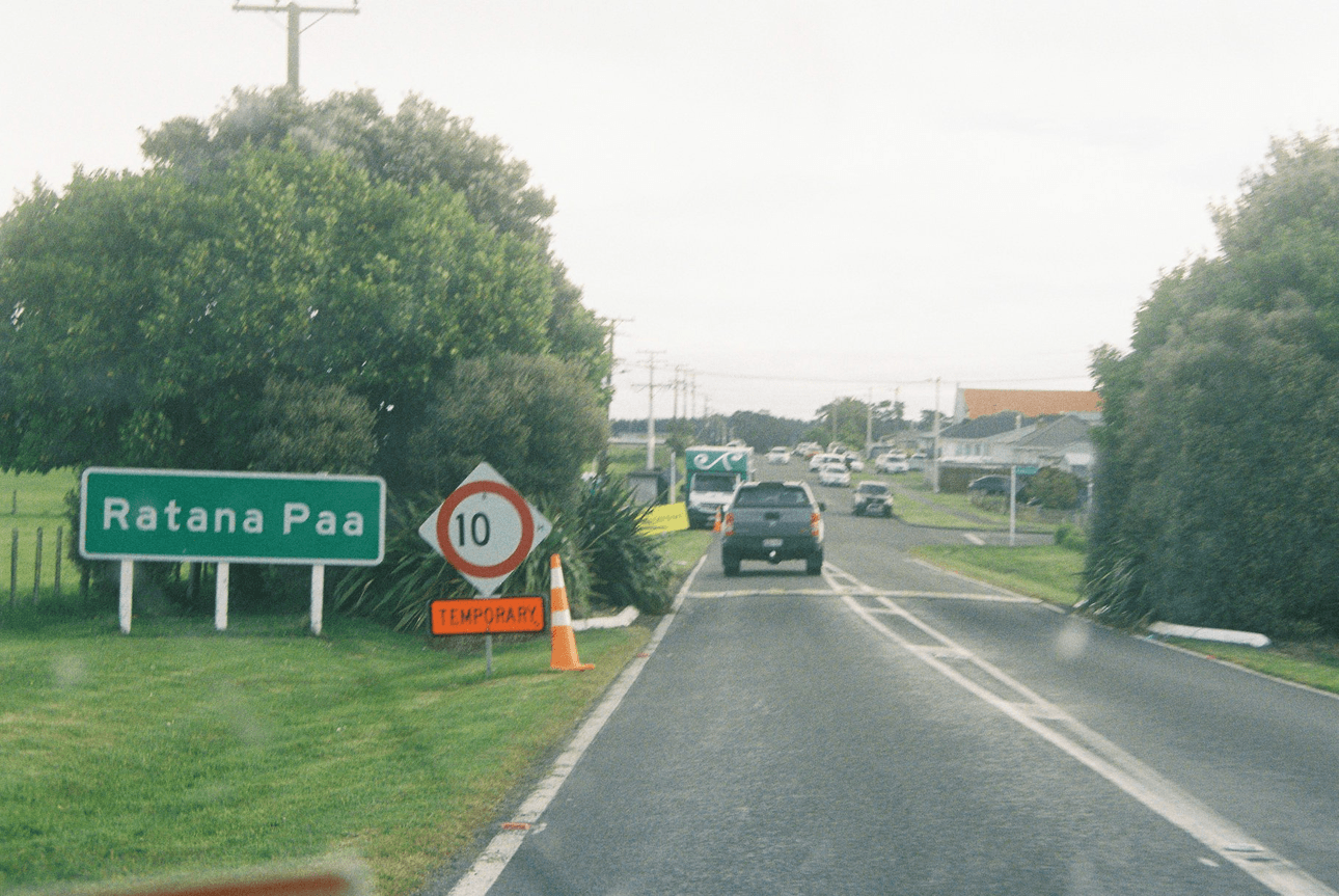 A road with a green sign reading "Ratana Paa," a speed limit sign of 10, and an orange "Temporary" sign. Cars are ahead in the distance, trees line both sides of the road, and houses are visible on the right.
