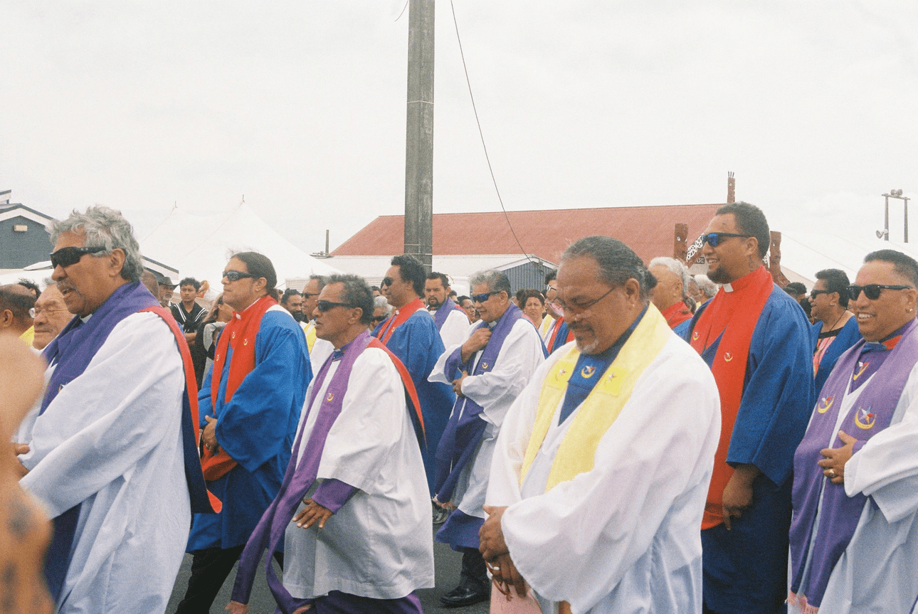 A group of people wearing colourful robes and sunglasses walk outdoors in a procession, some smiling. The robes are white, blue, red, and purple, and there are tents and a building in the background.