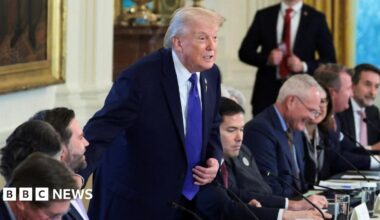 Donald Trump stands, one hand on his blue tie, at a table at which several other people are seated, in a formal room in the White House.