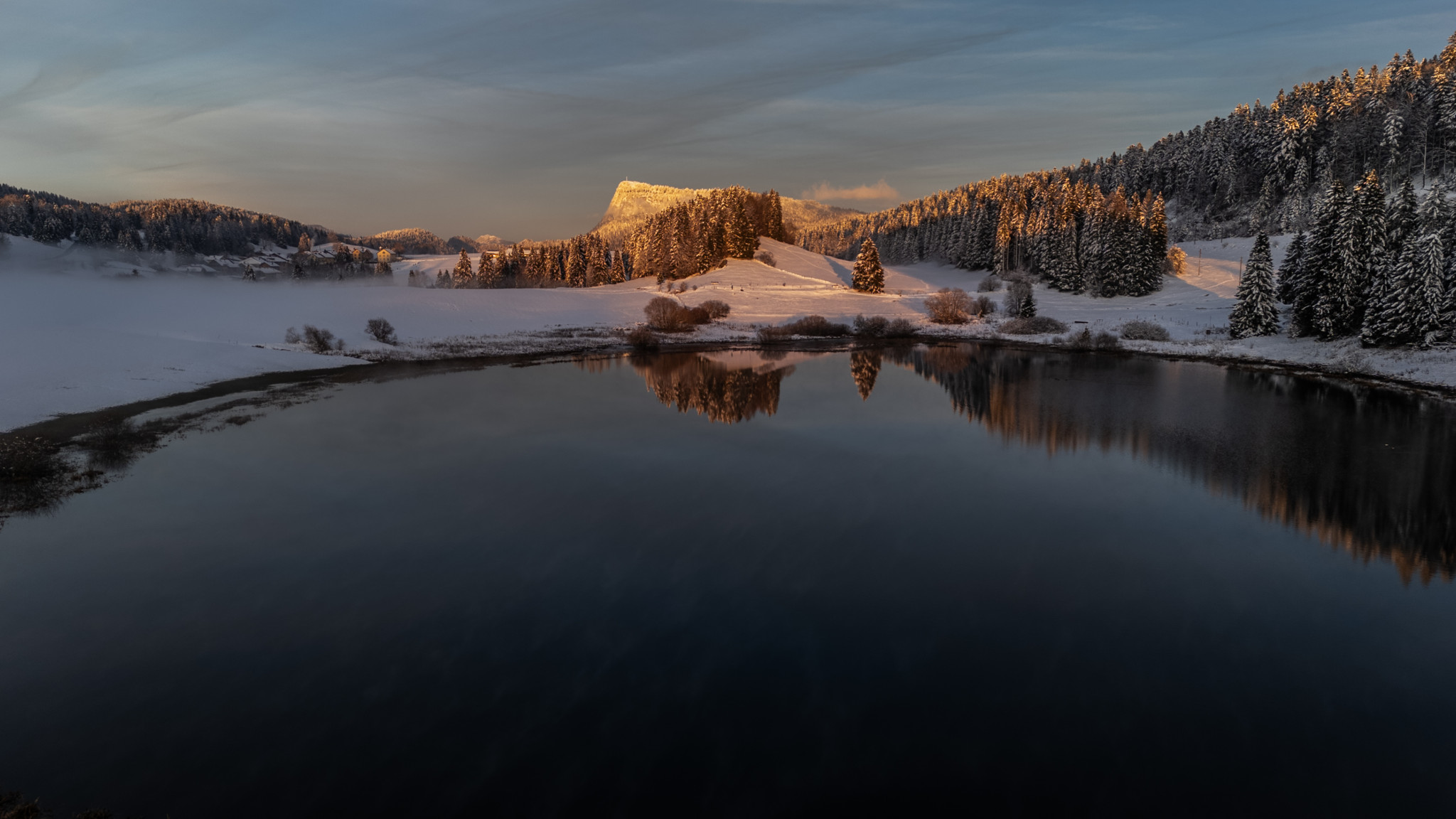 Paysage hivernal reflétant un lac calme entouré de forêts enneigées et de collines sous une lumière dorée.