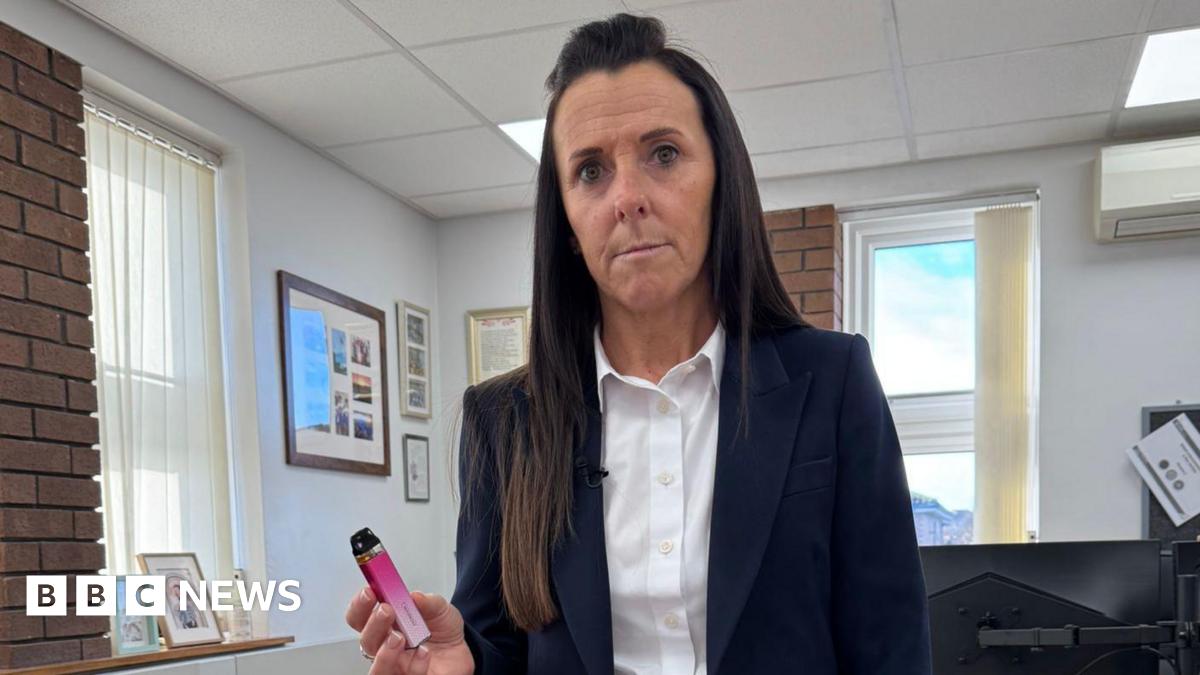 Head teacher wearing navy suit holds up a pink vape