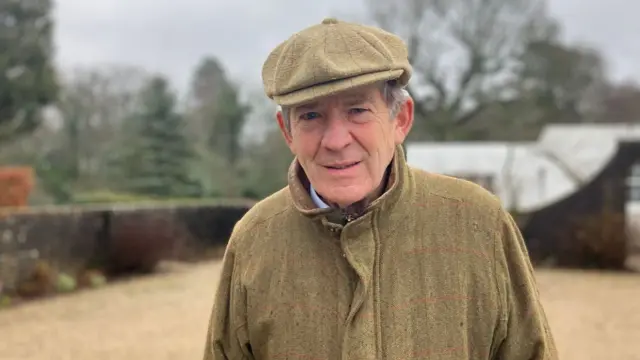 A man wearing a light green tweed coat and hat stood in a car park. Behind him is a brown brick wall and trees, though the background is blurred. He is elderly with grey hair coming out from beneath his hat. He is looking into the camera.