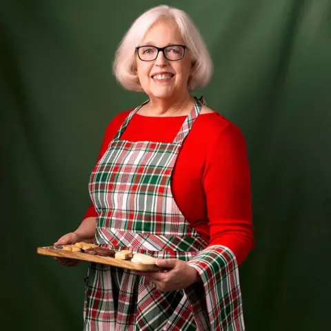 Scottish Gourmet USA Anne Robinson, who has grey hair and glasses, is wearing a tartan apron over a red top and is holding a tray of biscuits. She is smiling at the camera in front of a dark green background.
