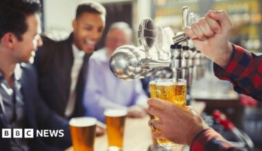 Close up of a pint being pulled from a tap. In the background there are blurred images of young men in shirts and jackets sitting at the bar with full pints in front of them. They look cheerful.