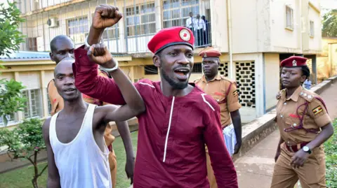 AFP via Getty Images Bobi Wine raises his fist in the air while handcuffed to another man. People wearing military-style outfits walk behing them.