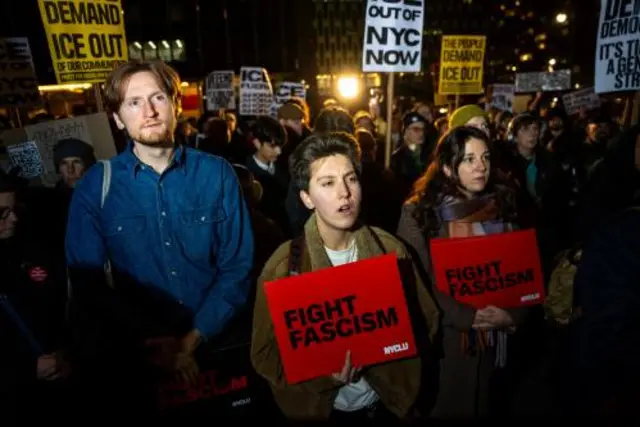 A man in a blue shirt and two women holding red signs stand in front of a crowd protesting ICE operations in Minneapolis