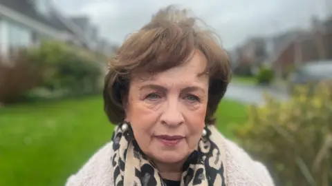 Diane Dodds is an older woman with short, brown hair. She is looking at the camera with a neutral expression on her face. She is wearing an animal print scarf and a furry, cream coat. She is standing outside a housing estate, but the background is blurred.