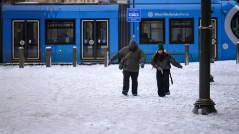 Getty Images Two people walk through slushy snow away from a tram labelled 'West Midlands Metro'