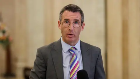 PA Media Muir speaking into a microphone. He is wearing a grey suit jacket, blue shirt and multi coloured striped tie. 