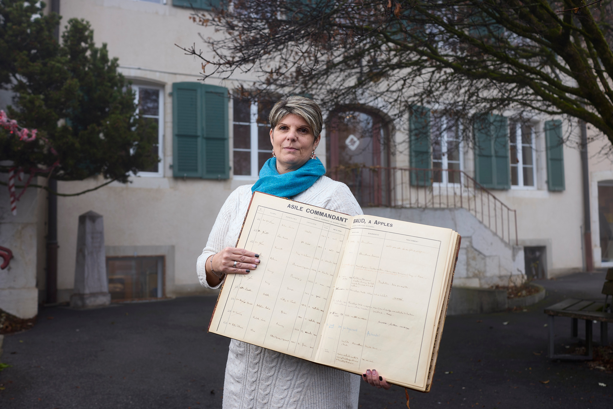 Anick Chevalley tient le premier registre des pensionnaires devant le bâtiment principal de l’EMS Fondation Baud à Apples, commémorant le fondateur Charles-Henri Alphonse Baud.