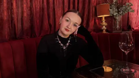 BBC A woman with a black jumper and decorative necklace sits at a table at a bar with a large empty wine glass. The sofa and curtains behind it are red